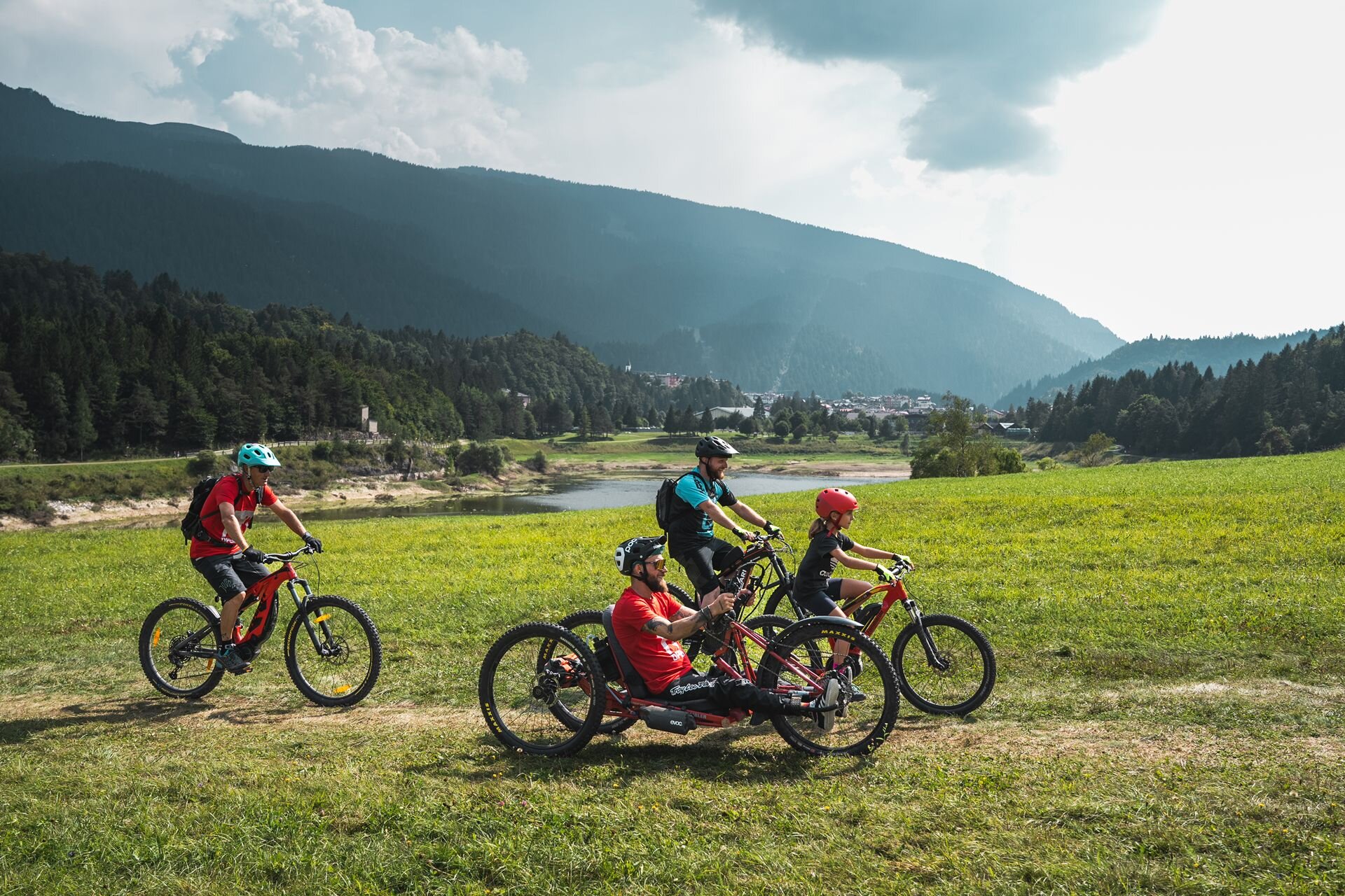 Un gruppo di ciclisti percorre un sentiero panoramico tra prati verdi e montagne. L’inclusività dello sport si riflette nella presenza di una handbike, rendendo l’esperienza accessibile a tutti. | © Camilla Pizzini, 2024