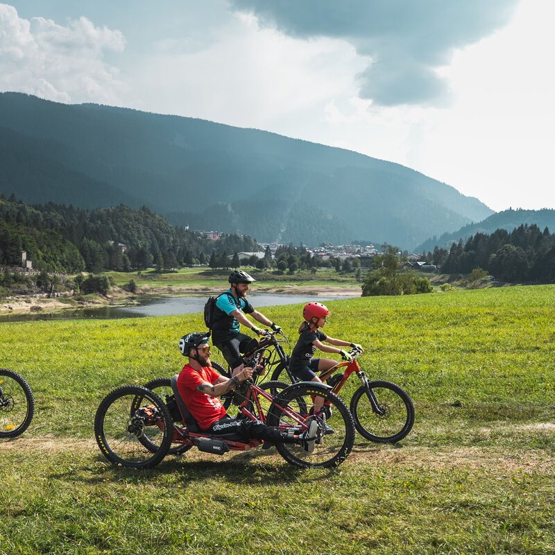 Un gruppo di ciclisti percorre un sentiero panoramico tra prati verdi e montagne. L’inclusività dello sport si riflette nella presenza di una handbike, rendendo l’esperienza accessibile a tutti. | © Camilla Pizzini, 2024