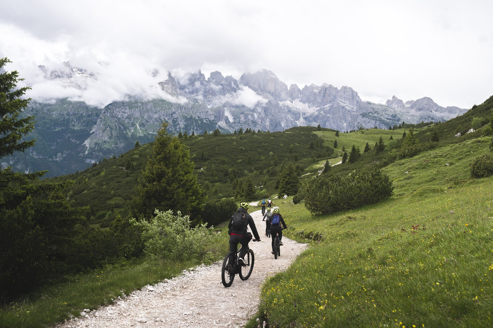 A group of five cyclists ride their e-MTB along a path on the Canfedin ridge. In the background, the Brenta Dolomites can be seen emerging through the fog. | © Marco Santinelli, 2022