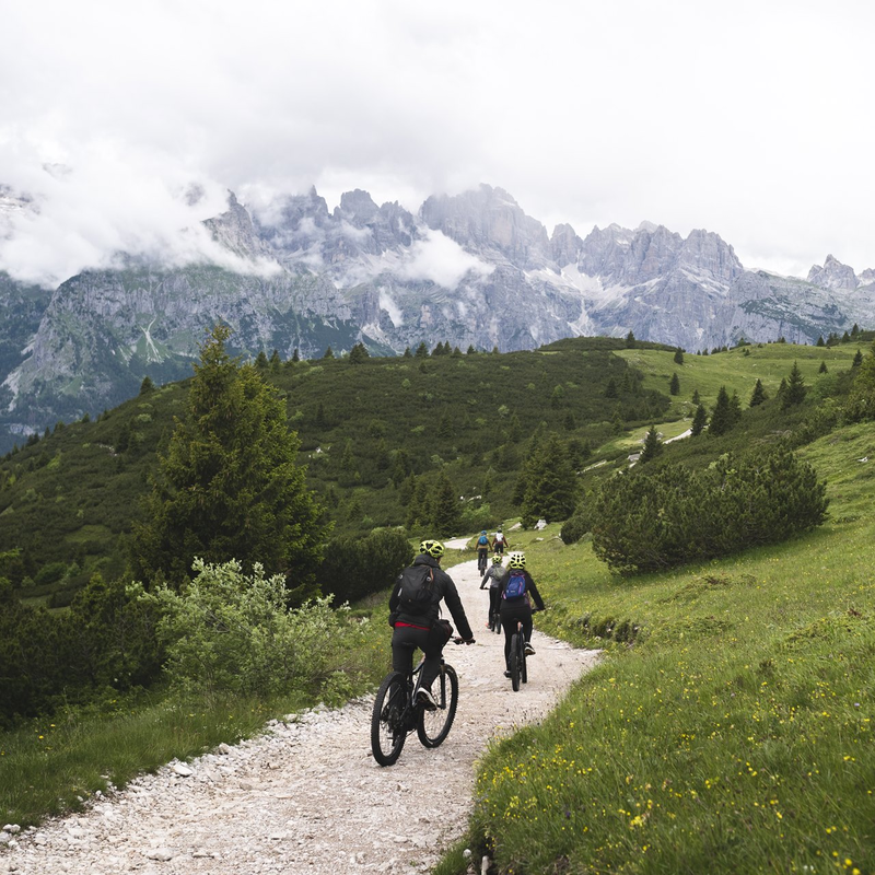 Un gruppo di cinque ciclisti percorre con l'e-MTB un sentiero sulla dorsale del Canfedin. Sullo sfondo si vedono le Dolomiti di Brenta che emergono tra le nebbie. | © Marco Santinelli, 2022