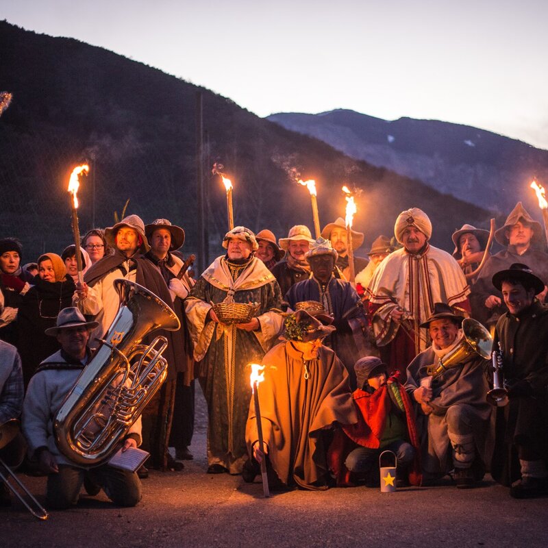 Un gruppo di persone in abiti tradizionali, alcuni con cappelli e mantelli, si è riunito all’aperto per celebrare la Canta della Stella. Alcuni tengono in mano torce accese, che illuminano i loro volti e creano un’atmosfera calda e suggestiva. Altri membri del gruppo suonano strumenti a fiato, come trombe e tube, mentre uno di loro tiene una grande stella decorata con bordi luccicanti, simbolo natalizio. Sullo sfondo si intravedono montagne scure sotto un cielo sfumato. | © Alfredo Croce, 2017