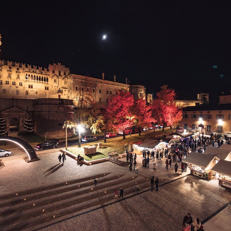 Questa immagine mostra il mercatino di Natale di Trento in notturna nella piazza antistante al Castello del Buonconsiglio. Il castello, illuminato da luci calde, ha una struttura imponente con una torre circolare sulla sinistra e mura merlate. Gli alberi lungo la piazza sono decorati con luci rosse che li fanno risaltare nell'oscurità. Le bancarelle del mercatino sono disposte in file ordinate, ognuna con un piccolo tetto bianco, e sono illuminate internamente, creando un’atmosfera accogliente e festosa. Diverse persone passeggiano tra le bancarelle, godendosi l'evento. Nel cielo notturno, una piccola luna è visibile sopra il castello. | © M. Gober - Archivio APT Trento