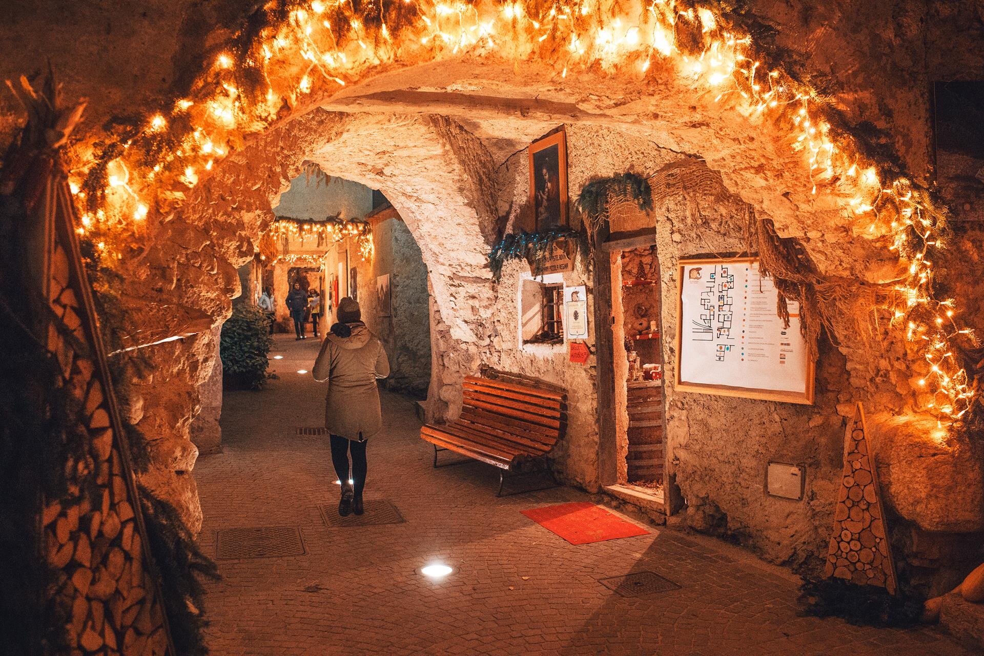 Un passaggio con pareti in pietra è decorato con luci natalizie calde e rami di abete. Una persona cammina lungo il corridoio, circondata da negozi artigianali, panchine e un'illuminazione diffusa proveniente da faretti sul pavimento. | © Filippo Frizzera, 2018