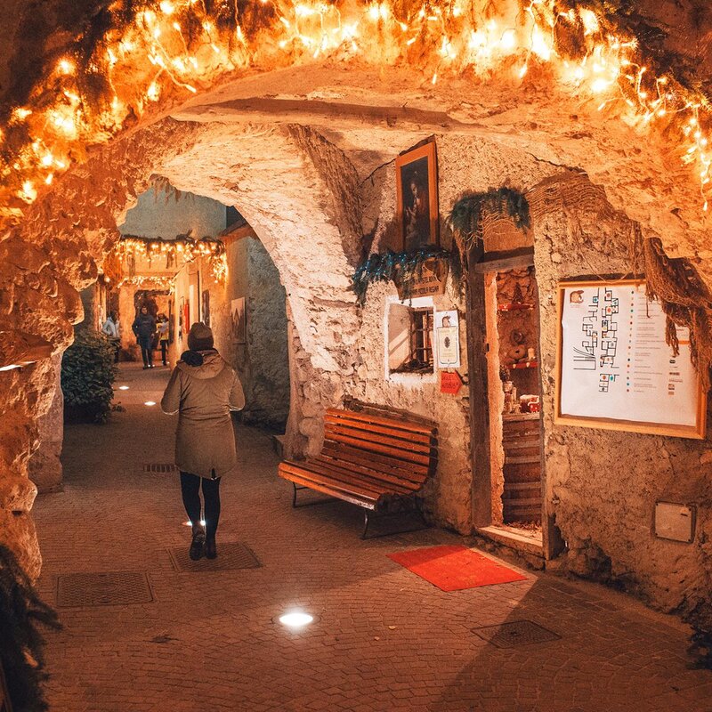 Un passaggio con pareti in pietra è decorato con luci natalizie calde e rami di abete. Una persona cammina lungo il corridoio, circondata da negozi artigianali, panchine e un'illuminazione diffusa proveniente da faretti sul pavimento. | © Filippo Frizzera, 2018