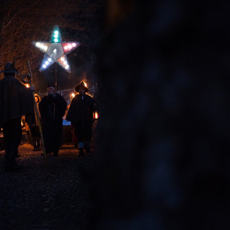 Un gruppo di persone avanza lungo un sentiero acciottolato durante la notte, portando fiaccole accese e una grande stella luminosa multicolore su un’asta. Sullo sfondo si intravedono alberi spogli, illuminati debolmente dalla luce delle torce. | © Alfredo Croce, 2017