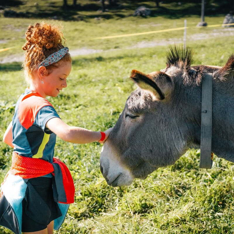 Una bambina con un completo da montagna e i capelli rossi accarezza il muso di un asino. | © Mirko Perli, 2023