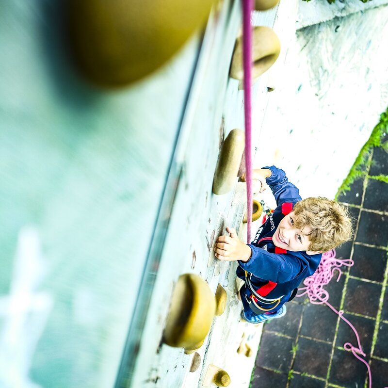 Un bambino guarda l'obiettivo mentre si arrampica sulla palestra di roccia all'Andalo Life | © Champion's Camp, 2018