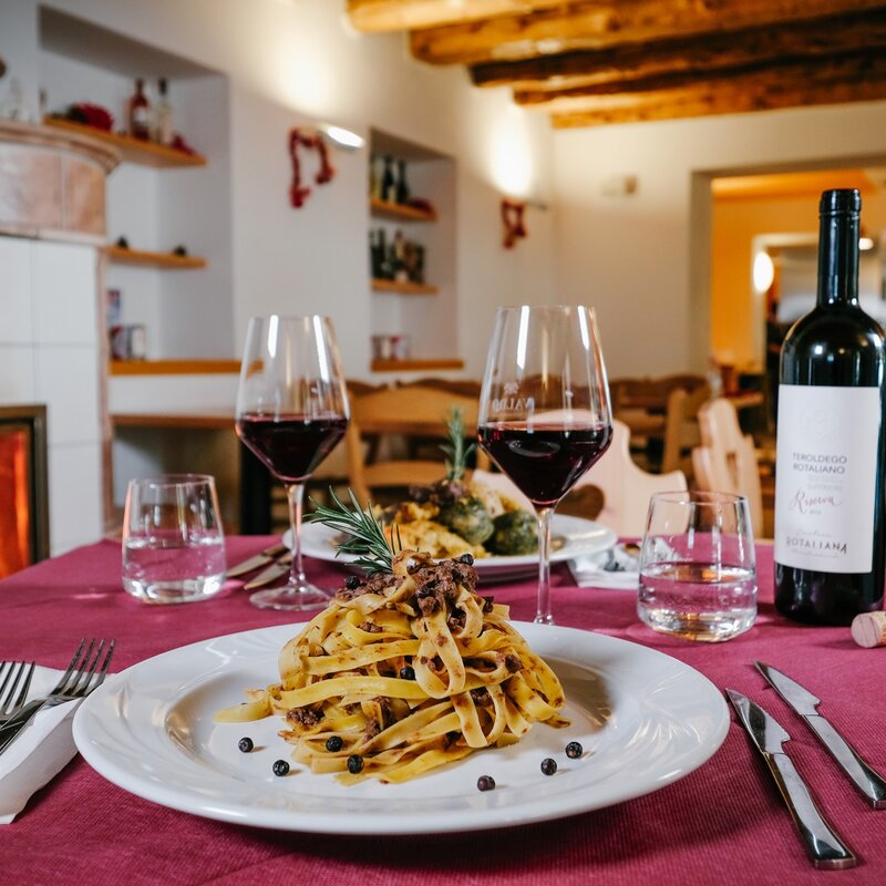 Un tavolo apparecchiato davanti al camino di Malga Terlaga. In primo piano un piattodi tagliatelle ai funghi e due calici di Teroldego Rotaliano, un vino rosso. Si intravede sullo sfondo un secondo piatto. | © Multiweb, 2019