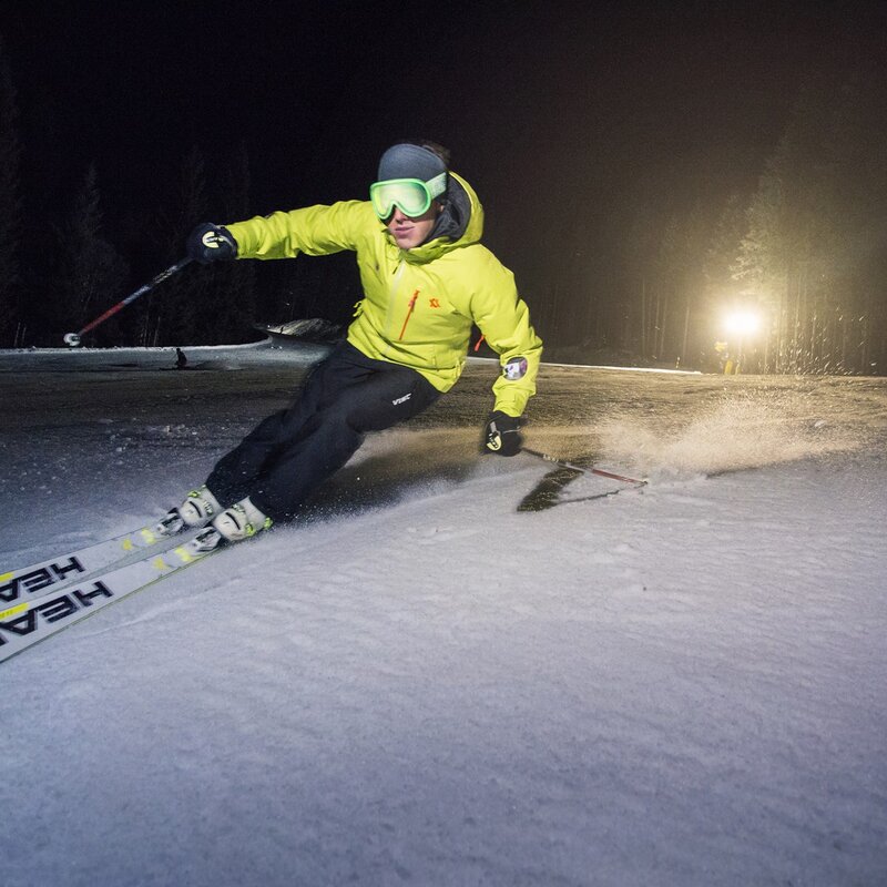 Questa foto mostra uno sciatore che scende una pista innevata di notte, indossando una giacca gialla, pantaloni scuri e occhiali da sci. L'illuminazione proviene da un potente faro alle sue spalle, che illumina la neve. Lo sciatore è in una posizione dinamica e inclinata. | © Filippo Frizzera, 2018