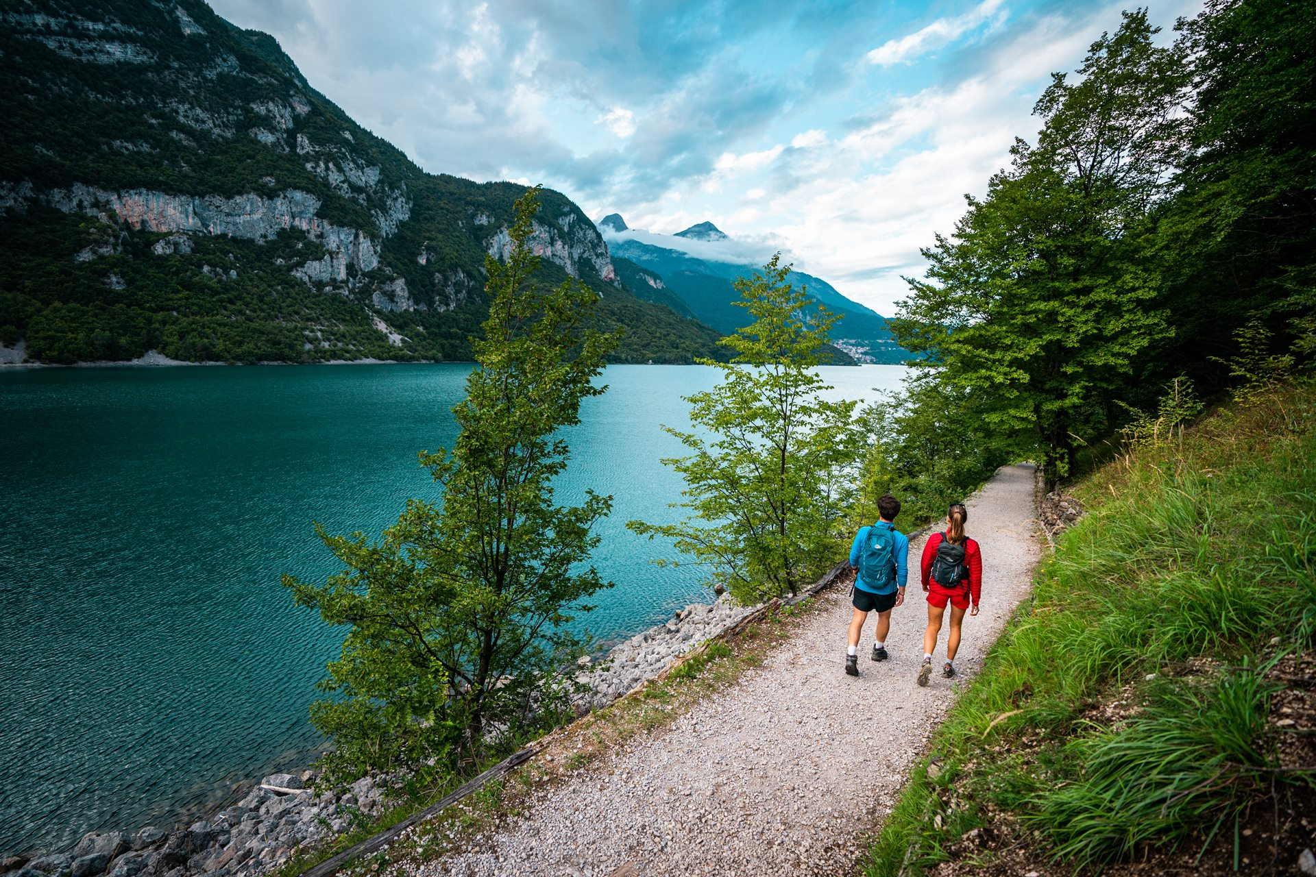 A couple walks along the Girolago path. On their left you can see Lake Molveno and the surrounding mountains. | © Camilla Pizzini, 2022