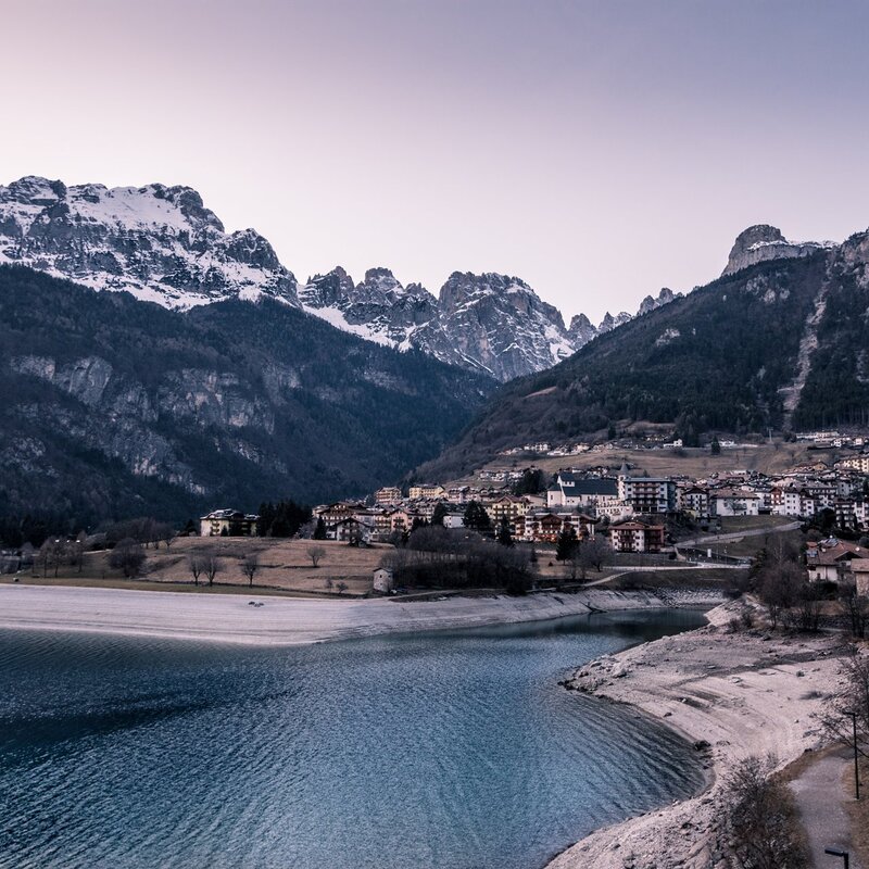 Un lago dalle acque calme e di un blu intenso occupa la parte inferiore dell'immagine, circondato da una spiaggia di sassi chiari. Sullo sfondo Molveno: case dai tetti scuri, edifici bassi e una chiesa visibile con il campanile. Dietro il paese, si ergono imponenti le Dolomiti di Brenta, coperte da foreste scure e da alcune cime innevate. Il cielo è limpido e sfuma verso toni chiari. Al tramonto l’atmosfera è serena e fredda, tipica di una giornata invernale. | © Benedetta Bernardeschi, 2020