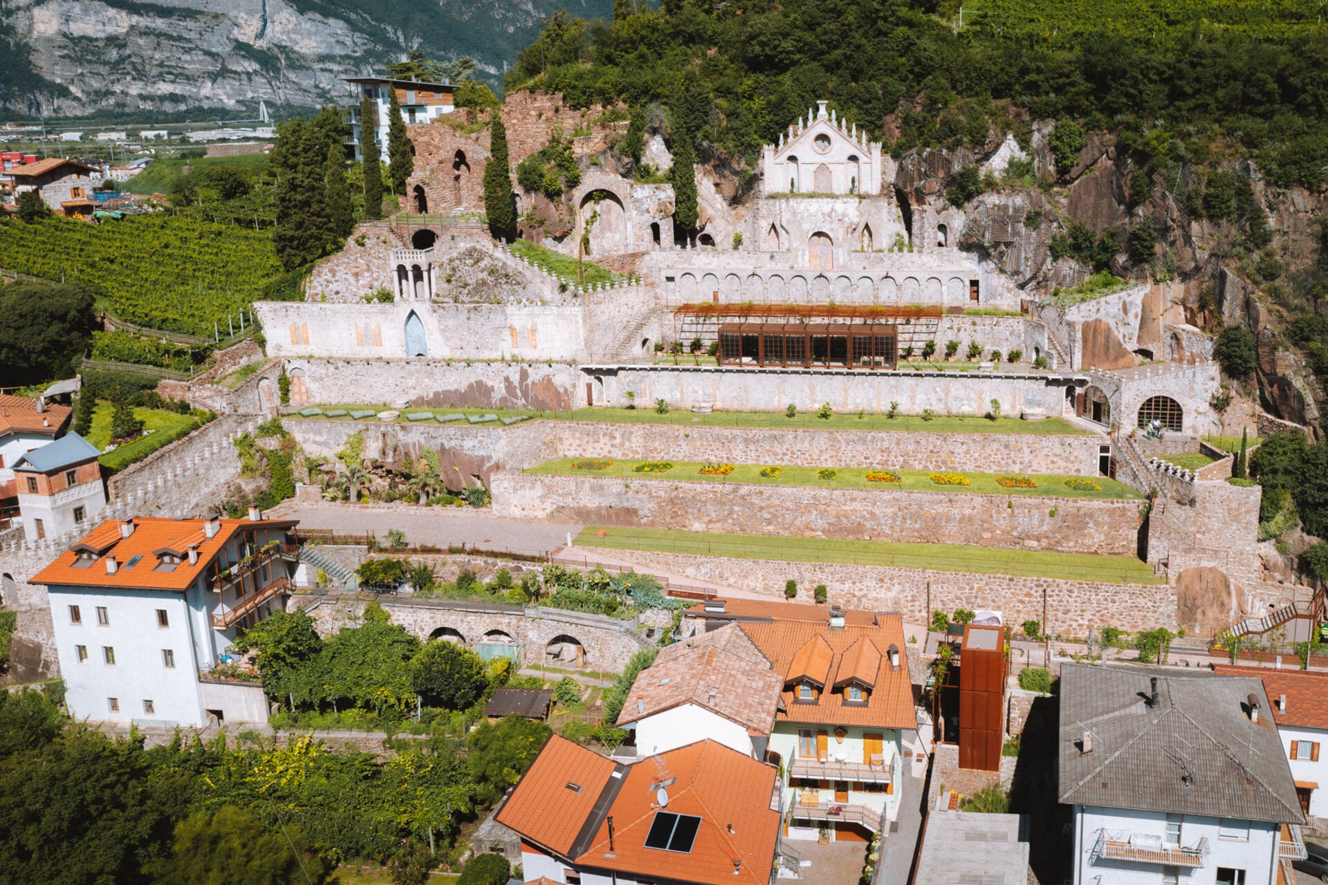 Ein antiker Komplex mit Steinmauern und einer mit Bögen und einer Rosette verzierten Fassade dominiert einen terrassenförmigen Hang mit gepflegten Gärten.  Am Fuße des Hügels überragen Häuser mit roten Dächern den Komplex und bilden einen Kontrast zwischen Antike und Moderne. | © Nicola Cagol, 2021