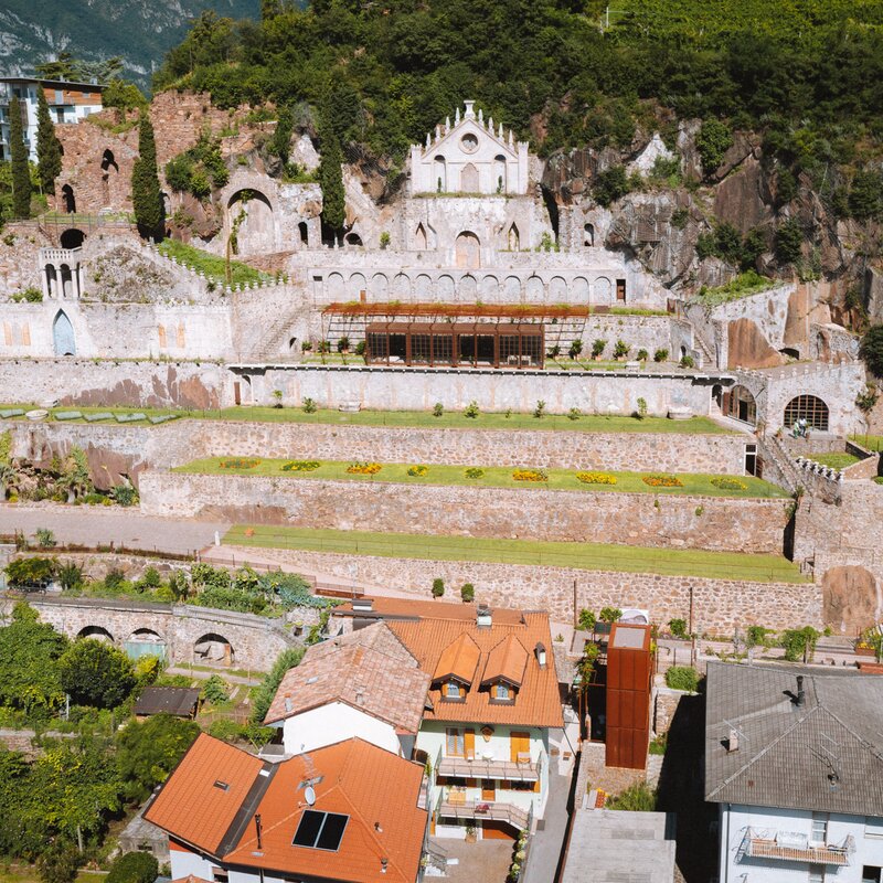 Un antico complesso, con mura in pietra e una facciata decorata con archi e un rosone, domina un pendio terrazzato con giardini curati.  Ai piedi della collina, case con tetti rossi si affacciano sul complesso, creando un contrasto tra antico e moderno. | © Nicola Cagol, 2021