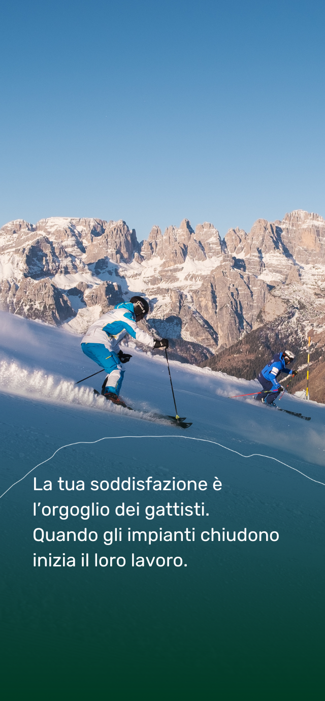 Nell'immagine: due sciatori scendono per le piste della Paganella con le Dolomiti di Brenta sullo sfondo. Nel testo: la tua soddisfazione è l'orgoglio dei gattisti. Quando gli impianti chiudono inizia il loro lavoro.  | © Filippo Frizzera, 2024