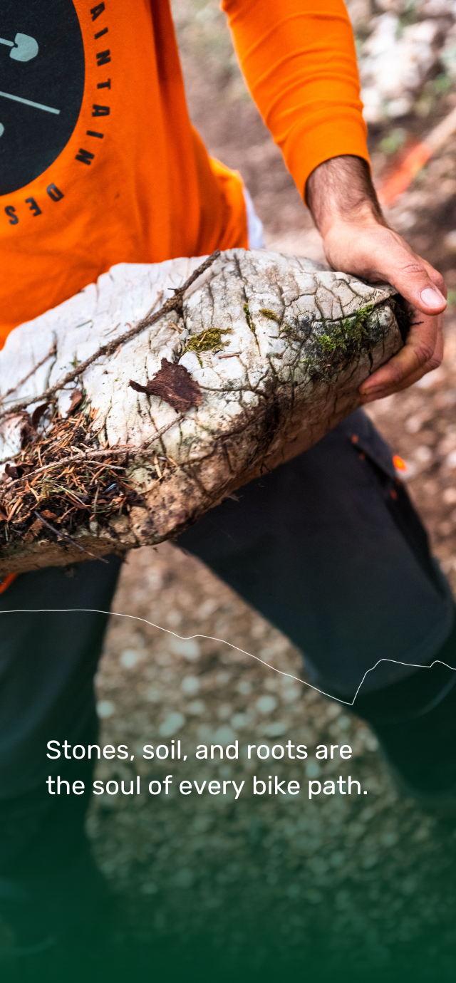 in the picture: a trail builders holds in his hands a large stone removed from the bottom of a mountain bike trail. in the text: Stones, soil, and roots are the soul of every bike path. | © Filippo Frizzera, 2024