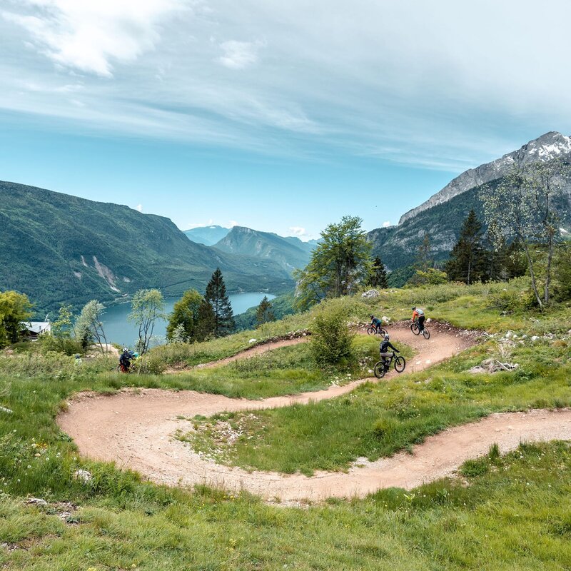 Biker fahren von der Big Hero-Strecke im Dolomiti Paganella Bike Park ab, mit Molveno im Hintergrund | © Giacomo Podetti, 2024